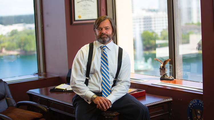 Dan Eckhart sitting on a large wooden desk in front of three windows